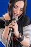 Placeholder: attractive female country music singer leaning forward while holding microphone in one hand, other hand brought up to side of neck, change clothing to plaid in natural tones, leather bracelets on wrists, long hair, mouth open singing, rings on fingers, eyes closed