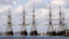 Placeholder: A seaport with several tall sailing ships docked, their masts and rigging visible against the sky. The ships appear to be from the 19th century, with wooden hulls and multiple masts. The scene suggests a busy maritime port from that historical period