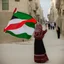 Placeholder: A very beautiful girl carrying a large Palestinian flag in her hands and waving it while wearing a keffiyeh and an embroidered Palestinian dress.