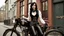 Placeholder: full-height portrait of a woman with straight shoulder-length black hair, with metal arms and legs, dressed in leather trousers, and a waistcoat, in a Victorian street next to a steampunk bike