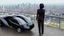 Placeholder: Tall thin woman, with straight black hair, dressed in a camouflaged jumpsuit, looking out from the rear of a futuristic aircar, on a tarmac runway, with a city skyline in the distance
