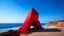 Placeholder: A prominent red V-shaped structure, possibly symbolic or artistic, rises boldly against a serene coastal backdrop. The monolithic figure is richly textured, evoking a feeling of raw, organic materialization, and contrasts markedly with the dull, earthy foreground. This foreground blends into the deep, ultramarine expanse of the ocean, which is placid beneath a softly lit sky. Subtle hints of ambient light create a shimmering effect behind the top of the "T," resembling an ethereal fog or cloud f