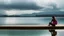 Placeholder: Woman sitting on a jetty with her feet in the water of the lake, in the background you can see a house of modern architecture that is reflected in the lake, the sky threatens storm