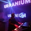 Placeholder: French cool synth player, he is called "Jean-Maurice Brûlée" performing at dance club, his name is "Jean-Maurice Brûlée" , white and blue Neon lights and smog, and "URANIUM" big sign on the wall, he is playing on synthesizers on stage with lot of laser beams and wearing black sunglasses, an earth is projected in background