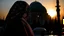 Placeholder: A Palestinian woman wearing an embroidered dress with the Dome of the Rock in front of her during sunset in winter.