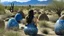 Placeholder: Outdoor landscape looking down. The foreground is an area of large blue and beige boulders of varying shapes and sizes. A tribal tattooed woman with long black hair is in the right corner. Tall bushes cover the ground, and distant mountains are in the background.