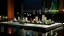 Placeholder: Architectural model floating on a dark, calm river at night, showing sleek metal vertical poles supporting minimalist white cubicles and vibrant spheres, with the city skyline in the background.