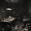 Placeholder: Warsaw, World War II. Photo of a dark, grey room. NO windows. A little bit destroyed and cluttered. There is a lot of things there. Brushes in cans with black paint on the floor. A giant table in the center. Above it there is a lightbulb. On the table there are a lot of papers. There is some solider boots in the corner, backpacks and a model of a truck car. We can see also a tiny photo of a young couple.