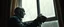 Placeholder: A photorealistic cinematic gothic horror scene of an extremely pale, sickly vampire quietly cleaning a large window in an ordinary old apartment during early morning. The vampire is doing the mundane task with calm, almost routine concentration, holding a worn cloth in one hand and a small spray bottle in the other. His skin is the main visual focus: extremely pale and unhealthy-looking, dry and uneven, with a naturally irregular organic texture, tiny pores, fine wrinkles, subtle flaky patches,