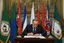 Placeholder: over the shoulder photo; politician signing a treaty; looking down; facing away, somber mood; in the background three banners, green banner, white banner, blue banner;