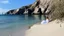 Placeholder: A serene coastal landscape featuring a sandy beach with intricate patterns, lush rocky cliffs in the background, and a calm sea with a distant sailing boat. A figure in a flowing white gown lies peacefully on the sand, surrounded by sparse vegetation. The sky is bright with soft clouds, evoking a sense of tranquility and dreaminess.