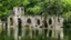 Placeholder: A ruined stone building partially submerged in a lake, balconies, verandas, arches, bridges, spires, stairs, trees, dense foliage, spanish moss, ivy, blue sky, white clouds