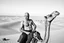 Placeholder: man looking like "Walter White" in breaking bad tv show, bald, with bare chest riding on a camel in sahara in desert, panorama, black and white photo, staring at a small town in horizon