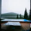 Placeholder: A polaroid photo of an odd room with structures. A transparent tarpaulin covers an image. There is a group of fir trees in the background and a high hill in the distance. A lake. The surface below is in a pastel color. Fog is present.