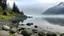 Placeholder: A wide shot of a lake with a rocky shoreline under a foggy sky. The lake is calm and reflects the sky, with some tall grass growing near the edge. A large pile of rocks forms a barrier or dam along the right side of the frame, sloping upwards. Several people are sitting or standing on the rocks near the water's edge. In the distance, mountains are visible through the fog. Streetlights line the top of the rocky barrier, disappearing into the fog. The color palette is muted, with shades of gray, b