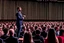 Placeholder: The image shows a confident speaker on stage, addressing a large audience with enthusiasm. The audience seems distracted and interested, and the setting exudes an atmosphere of elegance and impact.