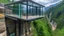 Placeholder: A house built on top of a rock, with panoramic glass walls that offer 360-degree views of a deep gorge and a wooden bridge that connects to the trail