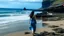 Placeholder: A young woman with long dark hair wearing a blue dress walking on a beach with rocky cliffs in the background and a calm, reflective ocean