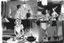 Placeholder: black and white storyboard, couple on foreground, we don t see them well because they are close to the camera but between them in the middle we see 3 chefs, scattered throughout the kitchen cooking, frying, cutting