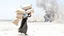 Placeholder: A Palestinian woman wearing a dress carrying very large bags of flour on her back, bending her back down in the destroyed Gaza City, and aid boxes descending from planes near the sea, with a large number of children looking up.