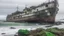 Placeholder: ABANDONED CARGO SHIP STRUCK ON THE COAST IN DETERIORATED CONDITION WITH RUST, ALGAE AND MARINE ORGANISMS, IT IS OBSERVED FROM ROCKS BY A PERSON, THE DAY IS VERY CLOUDY AND GRAY AND THE SEA IS ROUGH