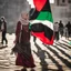 Placeholder: A very beautiful girl carrying a large Palestinian flag in her hands and waving it while wearing a keffiyeh and an embroidered Palestinian dress.