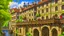 Placeholder: medieval buildings with balconies overhanging a river, blue sky and people, photorealism, trees, foliage, piers, intricate detail, ultra-sharp image, sharp focus