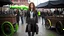 Placeholder: full-length portrait of a pale-faced woman with auburn wavy shoulder-length hair, with metal arms and legs, dressed in leather trousers and jacket, with a blouse, standing beside a steam carriage in a busy market square