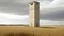 Placeholder: Aislamiento Funcionalista bajo Luz Nublada: Una vista frontal y clínica de una torre de elevador de grano de estuco beige pálido, similar a la de image_1.png, sola en un paisaje desolado de campos secos. El cielo es "Overcast" (nublado), proporcionando una luz uniforme y fría que resalta el desgaste de la pintura y las pequeñas ventanas cuadradas. Una solitaria farola de madera y una valla de alambre de espino en primer plano enfatizan el aislamiento. Fotografía de estilo documental de Bernd y H