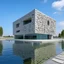 Placeholder: A large, modern, cuboid building with abstract-shaped cutouts stands centrally on a concrete slab that extends over calm, blue water. The building is made of a light gray material, possibly concrete, and its front facade is perforated with various organic, irregular holes, revealing glimpses of the interior and reflections of the sky and surroundings.