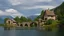 Placeholder: small gothic medieval house built into a rock face, lake, trees, arches, bridge, foliage, balconies, blue sky, white clouds