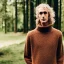 Placeholder: a full picture of a tall skinny man with blonde and frizzy hair wearing a woolen sweater standing outside in nature