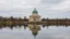 Placeholder: A classical-style building with a green dome stands on a platform extending into calm water. The building and surrounding trees are reflected in the water under a cloudy sky.