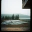 Placeholder: A polaroid photo of an odd room with structures. A transparent tarpaulin covers an image. There is a group of fir trees in the background and a high hill in the distance. A lake. The surface below is in a pastel color. Fog is present.