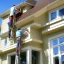 Placeholder: Two dudes standing on a ladder reaching up onto the edge of a house installing seamless gutters to the fascia