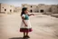 Placeholder: A five-year-old Palestinian girl wearing a traditional dress and new shoes looks to the side and points at a distant building.