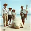 Placeholder: 1930's washed out faded color photography of European explorers dressed in kakhi pants and clean pressed white button-down shirts and safari hats posing with an Samoan native guide on a beach, giant eely sea creature washed up on beach in front of them