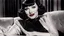 Placeholder: Claudette Colbert, dressed in stockings and stilettos, with a black bob hairstyle with bangs and dark sultry eyes and red lips, lying on a couch, smiling.