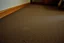 Placeholder: cheap motel room floor, worn carpet, very faint uneven discoloration in carpet, barely noticeable, irregular edges, subtle variation in tone, harsh flash lighting, poorly framed, candid documentary photo, not centered, no clear subject