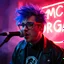 Placeholder: A MC performing at dance club, his name MC DRAGAN and looks crazy, crazy blue hair with Elvis Costello glasses. Neon lights and smog, MC DRAGAN SIGN ON WALL
