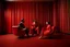 Placeholder: Un hombre vestido con traje, un hombre con pijama rojo, y una mujer vestida con traje, sentados en sillones, en una habitación de suelo de baldosas con líneas zigzagueantes, la habitación la recorre una gran cortina roja, fotografía realizada con cámara Leica y objetivo de 50 mm