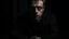 Placeholder: In the photo, we see a man sitting in the dark, looking at the camera with a cold gaze. The man is dressed in black and seems uninterested in what is going on around him.