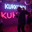 Placeholder: MR Kukrunk is Benny and DJ Roy in a disco, dancing, "Kukrunk" on sign wall, disco light, strobe, disco ball, dancing people