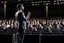 Placeholder: The image shows a confident speaker on stage, addressing a large audience with enthusiasm. The audience seems distracted and interested, and the setting exudes an atmosphere of elegance and impact.
