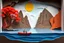 Placeholder: generate layered paper art that creates a beautiful dimensional landscape scene. The artwork uses a technique called paper cutting or paper sculpture, where multiple layers of colored paper are precisely cut and stacked to create depth and a three-dimensional effect. The scene depicts a serene moment with a lone figure standing by a canoe on calm water, surrounded by towering rock formations or canyon walls. The most eye-catching element is the vibrant red-orange tree on the left, which appears