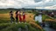 Placeholder: a group of young ladies in sports pants and blouse are dancing to camera in village over high grassy hills,a small fall and river and wild flowers at river sides, trees houses ,next to Ripe wheat ready for harvest farm,windmill ,a pretty train is arriving to station along river,a few village local shops ,cloudy sun set sky