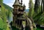 Placeholder: small wooden medieval gothic twisted house built into a cliff, trees, balconies, walkways, bridge, sunshine, lake