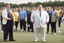 Placeholder: Overweight white politician wearing a suit; next to him a group of thin, muscular athletes in sports clothes; outside, sports field; realistic, photographic