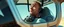 Placeholder: close up on weird out expression of guy suspended in straps inside laid back futuristic sci-fi glass human form car pillar on wheels, and a tracked wheel in the middle of the car