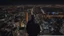 Placeholder: An Englishman in a bomber jacket standing at the top of a tall building looking across a city at night